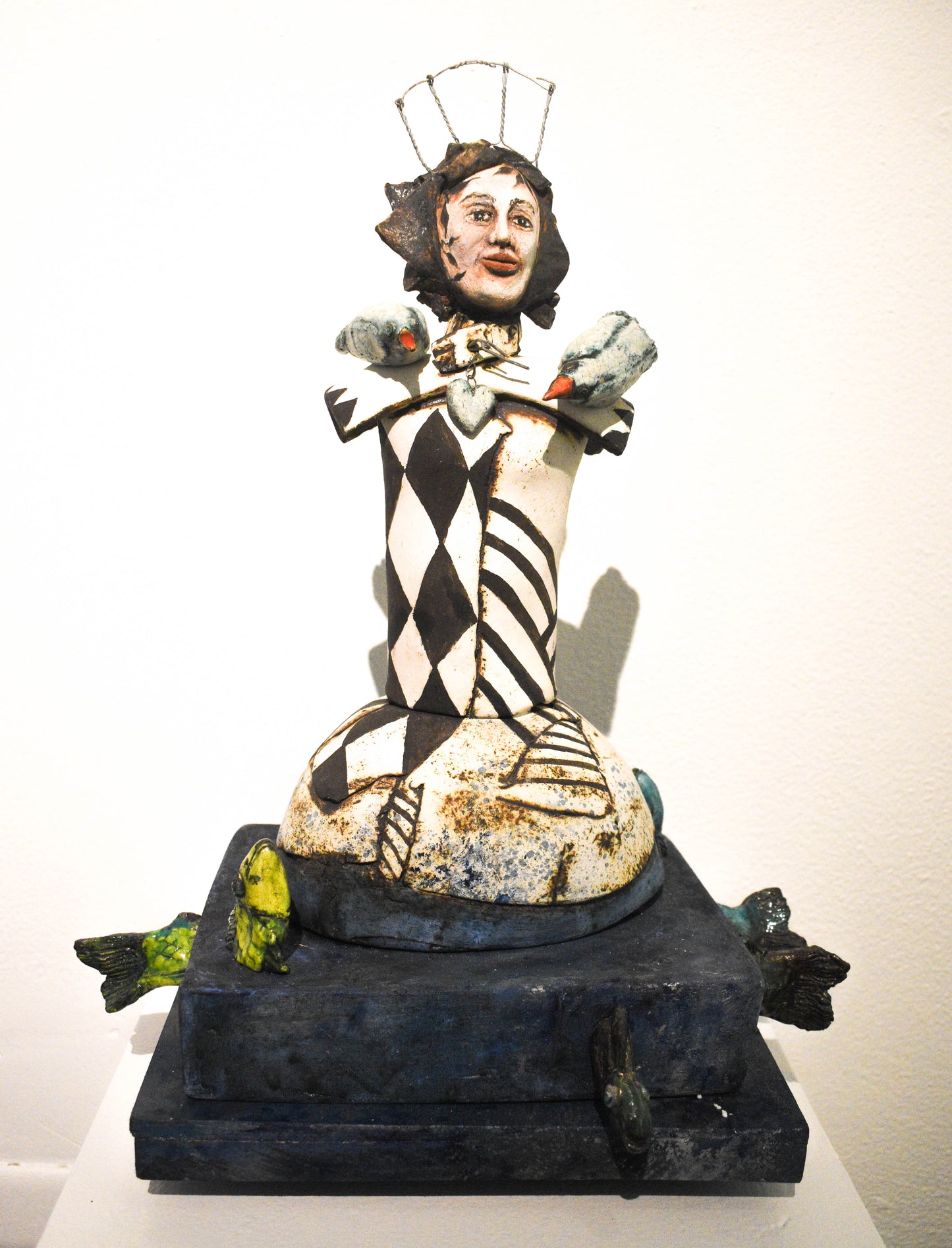 A ceramic sculpture featuring a stylized, abstract figure with geometric patterns in black and white, seated atop a textured base with green accents and perched birds.