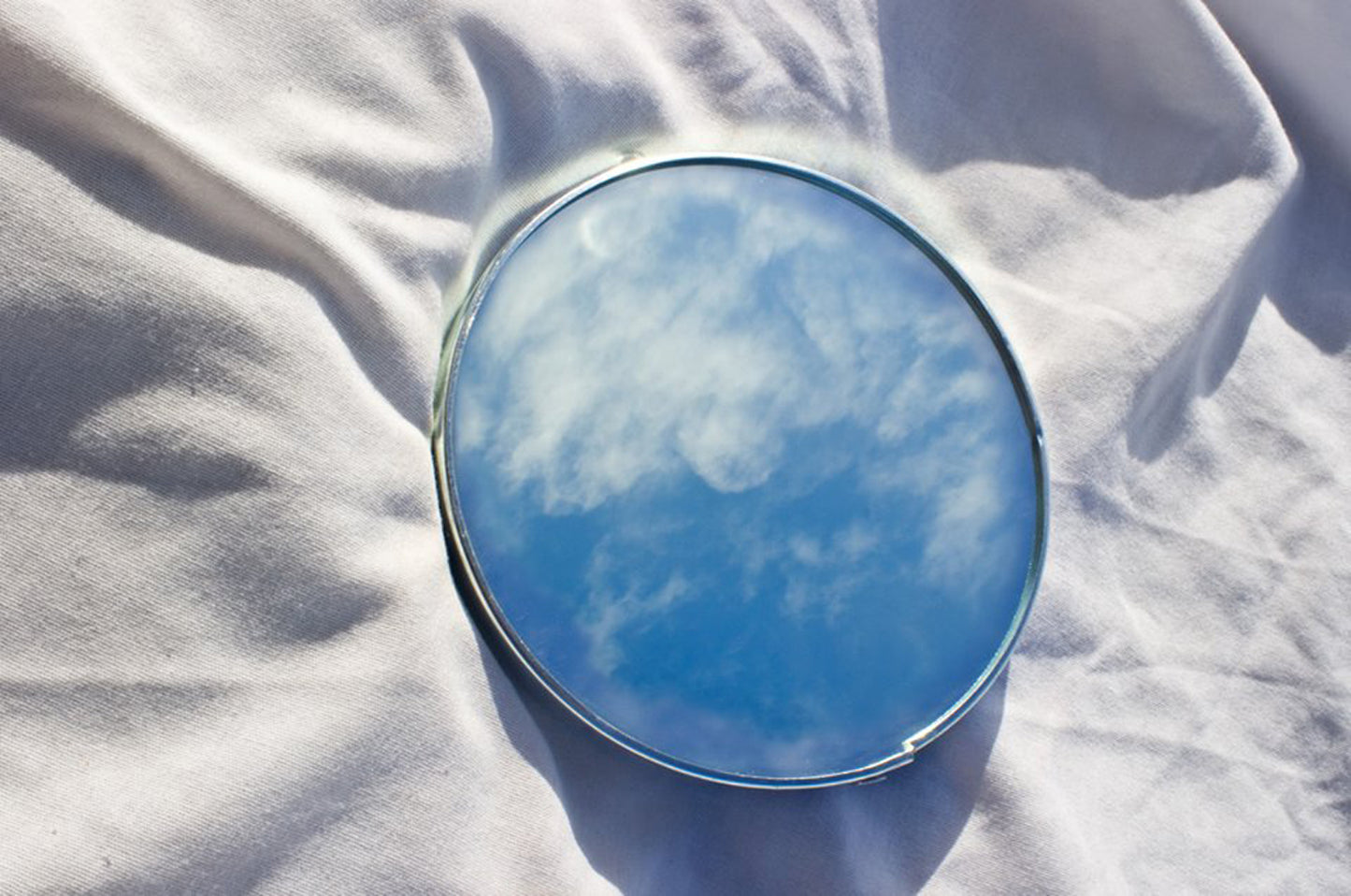 A round mirror laying on a cloth reflecting a cloudy sky.