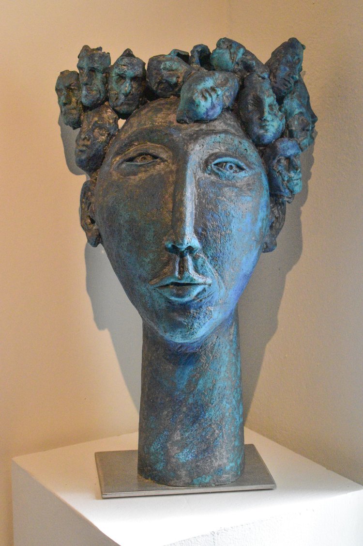 A bronze ceramic sculpture of a face with many small faces crowning it's head, with a stainless steel base, displayed on a shelf.