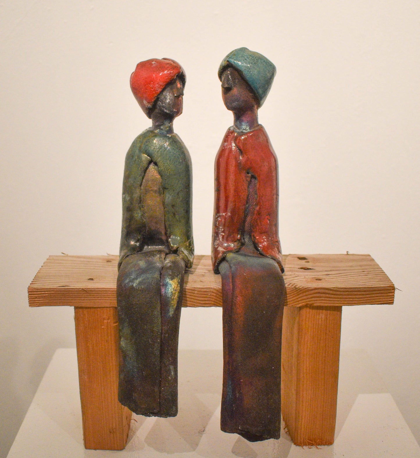two ceramic figures sit slightly facing each other sitting on wooden bench