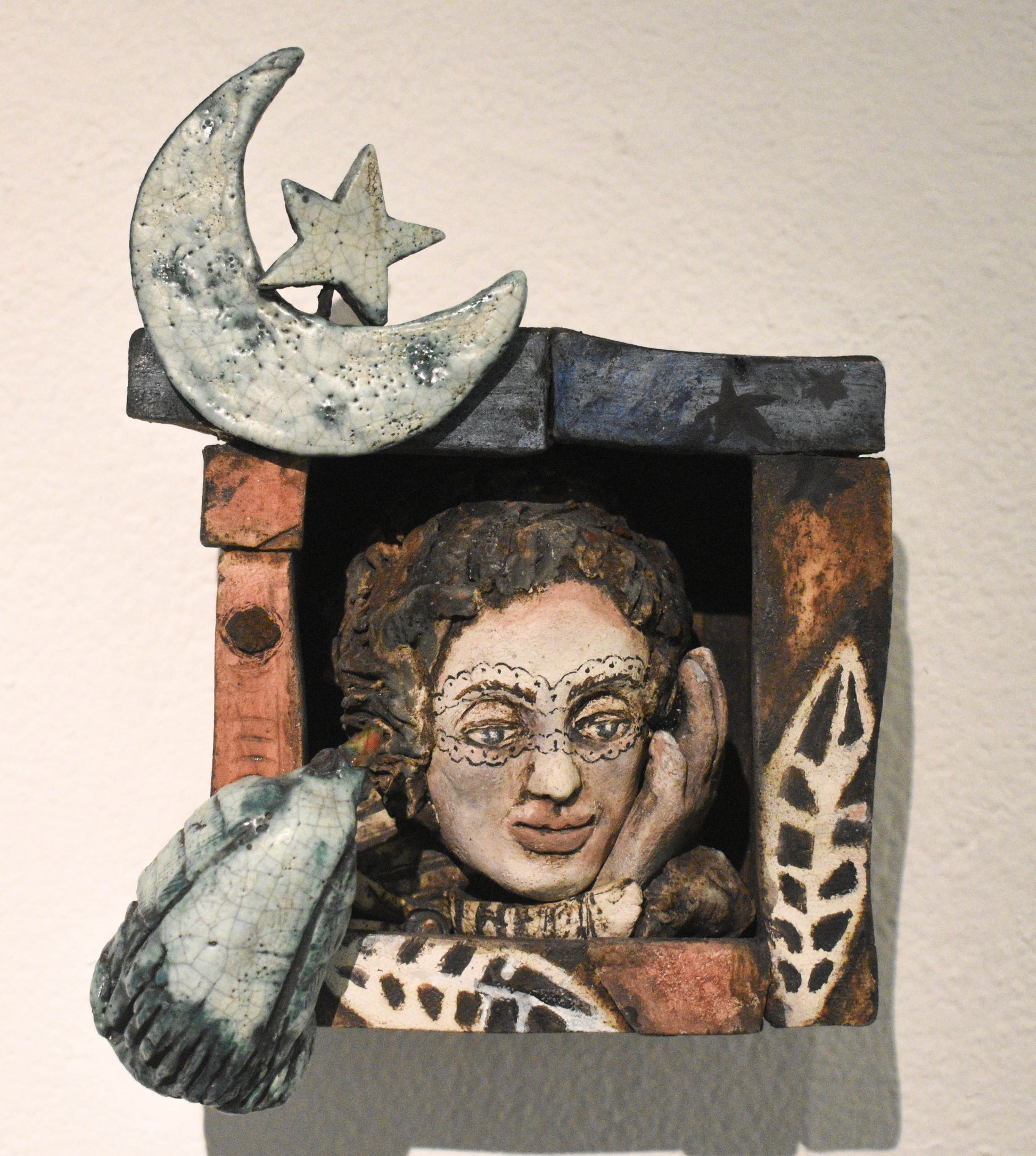 A small ceramic sculpture featuring a whimsical face with glasses, surrounded by abstract shapes and a crescent moon with a star on top.
