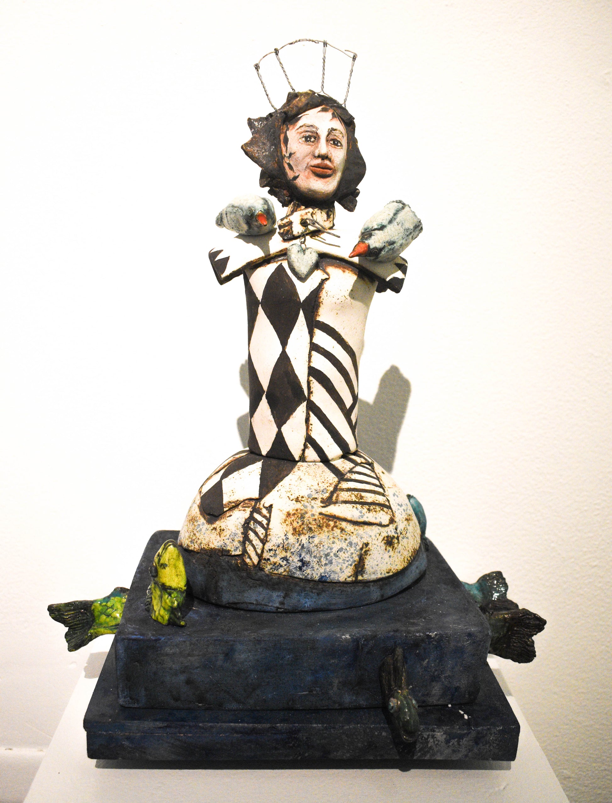 A ceramic sculpture featuring a stylized, abstract figure with geometric patterns in black and white, seated atop a textured base with green accents and perched birds.