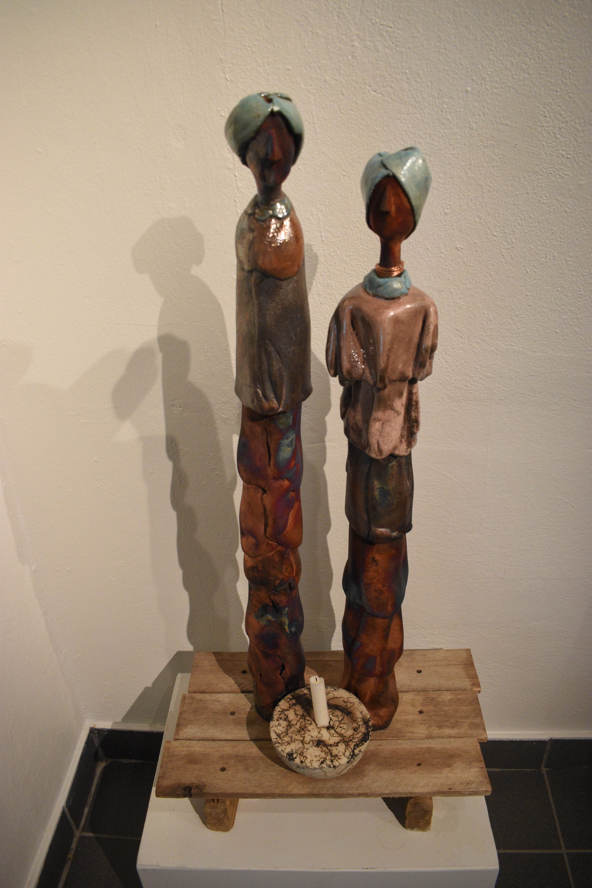 Two ceramic figures on a wooden platform, one figure is slightly in front of the other. The figures appear to be a man and a woman, with the man standing behind the woman, both wearing traditional clothing.